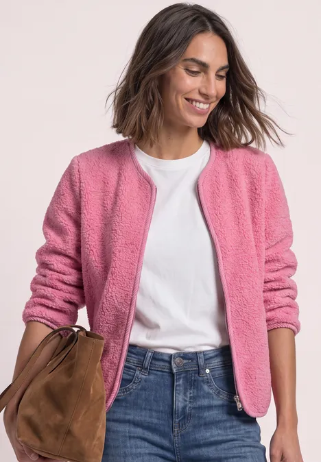 Eine lächelnde Frau in rosa Jacke, weißem T-Shirt, blauen Jeans mit brauner Tasche.