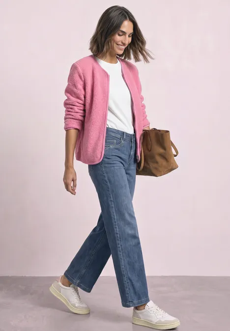 Eine Frau in weißem Shirt, pinkem Cardigan, blauen Jeans und weißen Sneakern trägt eine braune Tasche.