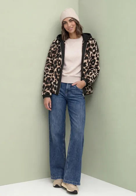 Teddy-Blouson mit abnehmbarer Kapuze Black Frau trägt Beanie, cremefarbenen Pullover, Leopardenjacke, weite Jeans und beige Stiefel vor neutralem Hintergrund.