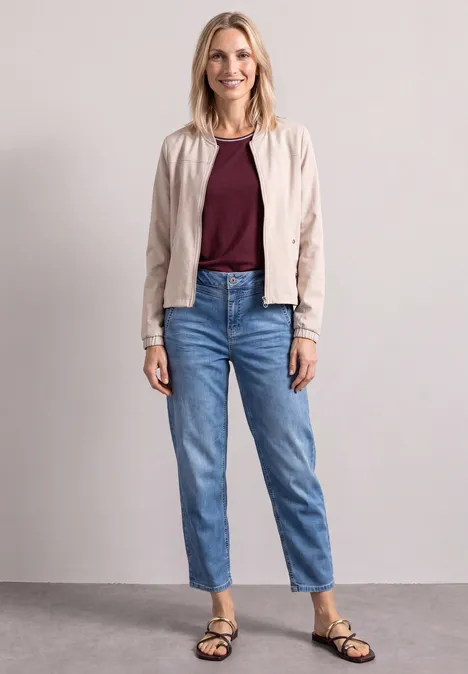 Frau in Jeans, bordeauxrotem Top, heller Jacke und Sandalen vor neutralem Hintergrund.