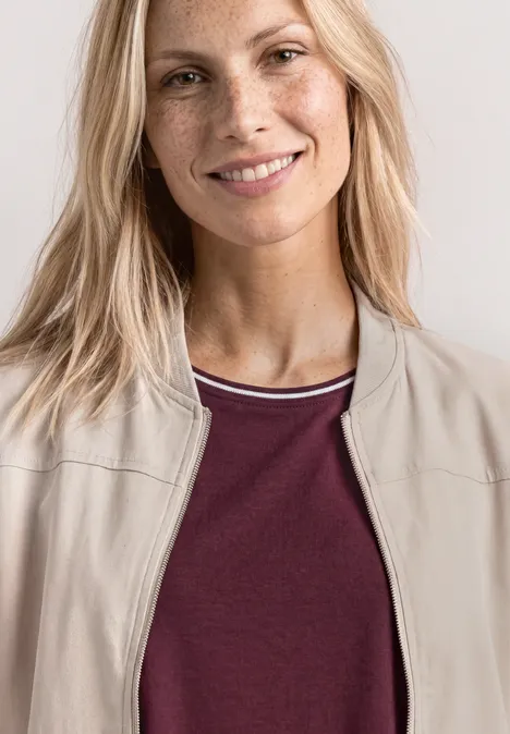 Blonde Person von den Schultern aufwärts, trägt eine beige Jacke und ein bordeauxrotes Shirt.