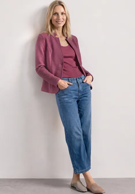 Vrouw leunt tegen witte muur, draagt paarse blazer en top, blauwe jeans en beige ballerina's.