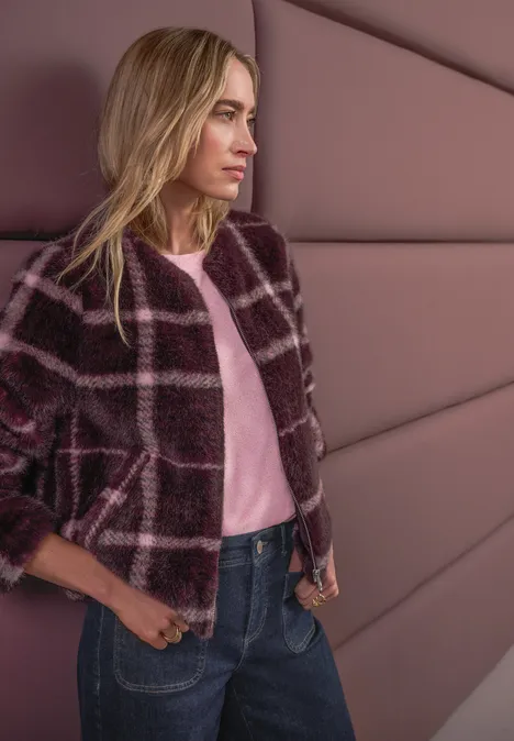 Dame mit kariertem Mantel lehnt an gepolsterter Wand, trägt rosa Shirt und Jeans.