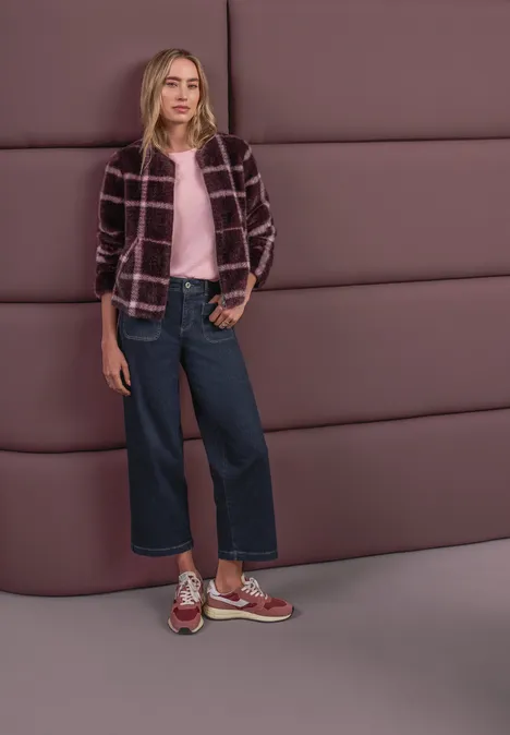 Frau trägt rosa Pullover, karierte Jacke, weite Jeans und Sneakers vor gepolsterter Wand.