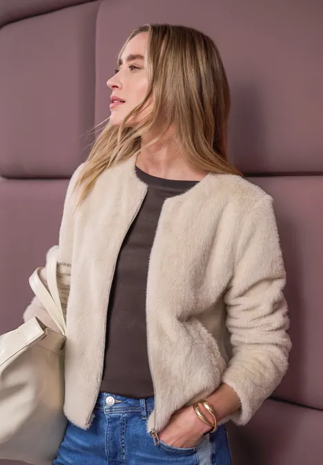 Frau in beiger Teddyjacke, dunklem Top und Jeans mit Tasche vor purpurfarbener Wand.