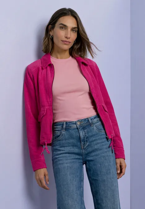 Une femme en veste rose, top rose et jean devant un mur violet.