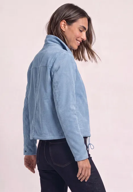 Femme de profil portant une veste en velours côtelé bleu clair et un jean foncé sur fond neutre.