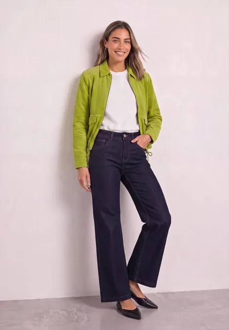 Frau lehnt an Wand, trägt grünen Blazer, weißes Shirt, dunkle weite Hose und schwarze Ballerinas, lächelnd.