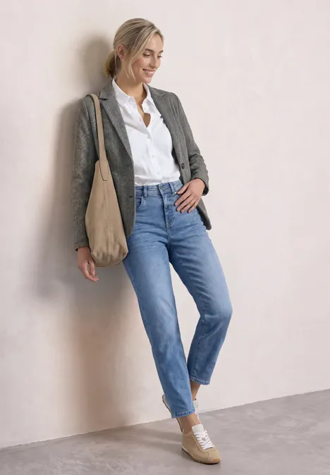 Frau in weißer Bluse, grauem Blazer, Jeans und Tasche steht lässig an Wand.
