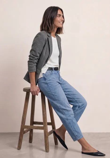 Femme souriante assise sur un tabouret, vêtue d'un blazer gris, top blanc, jean bleu et ballerines noires.