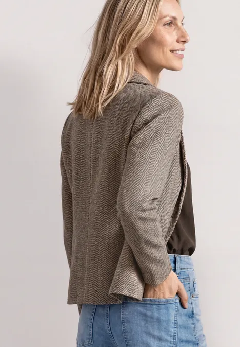 Seitenansicht einer lächelnden Frau in grauem Blazer und Jeans, Hand in Tasche vor hellem Hintergrund.
