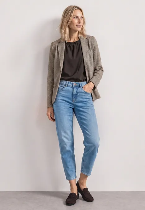 Eine Frau mit blondem Haar trägt grauen Blazer, schwarzes Shirt, blaue Jeans und schwarze Slipper.