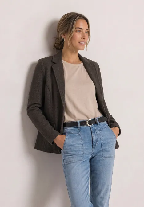 Uśmiechnięta kobieta w casualowej stylizacji: marynarka, t-shirt i jeansy, oparta o jasną ścianę.