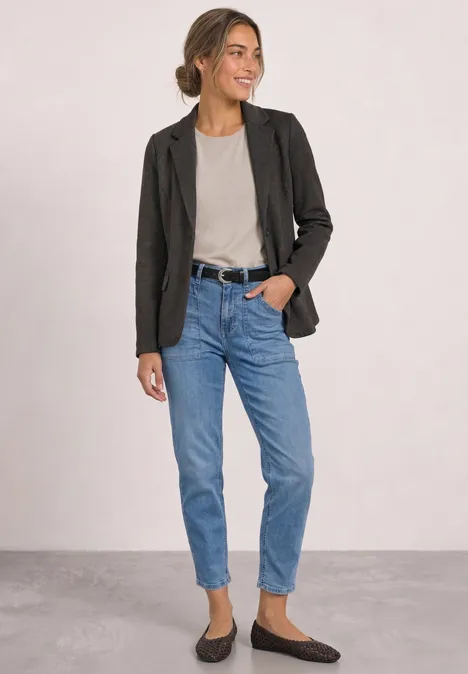 Frau in schwarzem Blazer, grauem Shirt, blauen Jeans und braunen Loafers, lächelnd seitlich stehend.