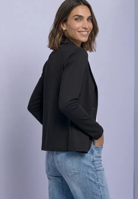 Eine Frau in schwarzem Blazer und blauen Jeans steht seitlich lächelnd vor hellem Hintergrund.