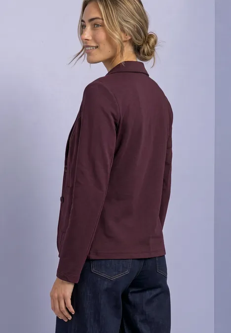 Eenvoudige blazer in effen kleur rustic rouge Een vrouw in een bordeaux colbert en spijkerbroek kijkt over haar schouder, tegen een lila achtergrond.