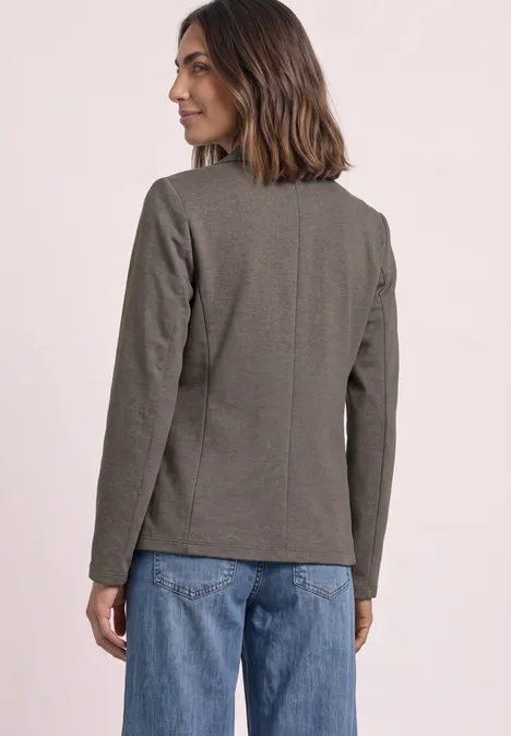 Frau steht seitlich gewandt, trägt graues Sakko und blaue Jeans vor hellem Hintergrund.