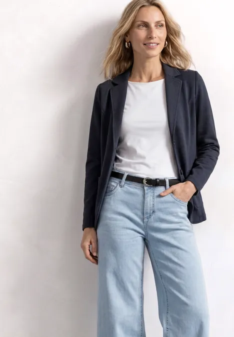 Frau in weißem T-Shirt, dunkelblauer Jacke, hellblauen Jeans und schwarzem Gürtel vor hellem Hintergrund.
