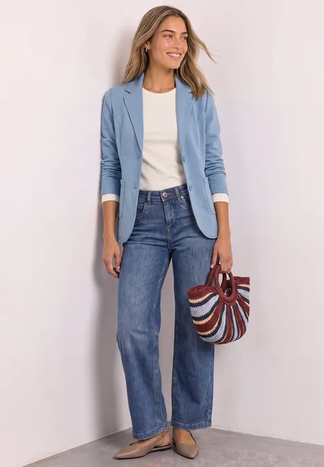 Vrouw in lichtblauwe blazer, wit shirt, wijde jeans en gestreepte tas voor een neutrale achtergrond.