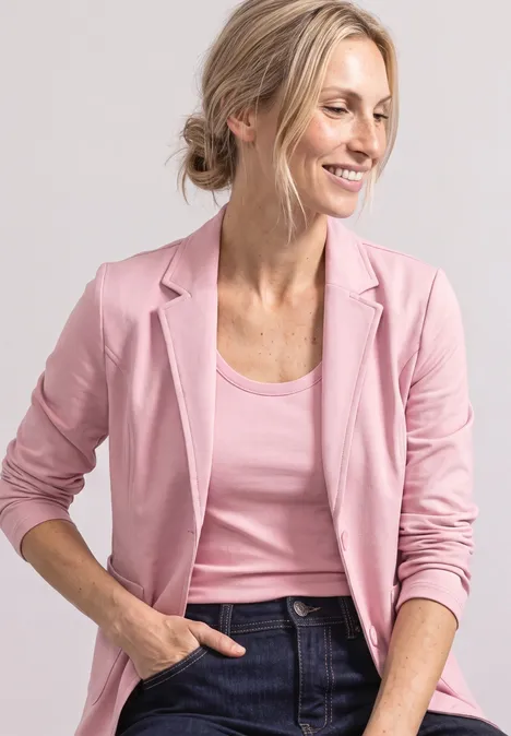 Lächelnde Frau in rosa Blazer und Top, mit Jeans und Hand in der Tasche.