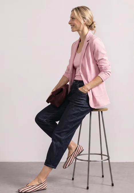 Eine stilvolle Frau sitzt auf einem Hocker, trägt rosa Blazer, Jeans und gestreifte Ballerinas, hält eine Clutch.