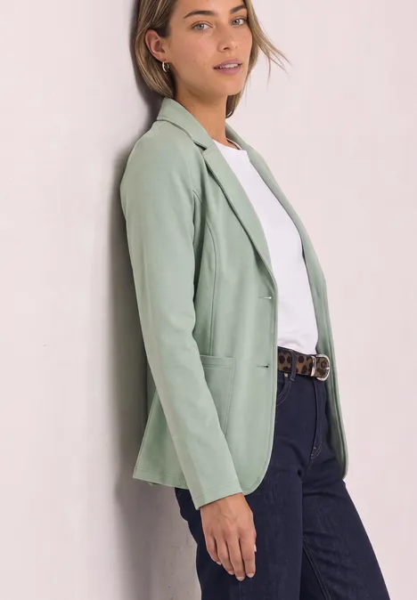 Frau lehnt an Wand, trägt mintgrünen Blazer, weißes Shirt, blaue Jeans und Gürtel.