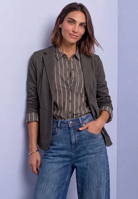 Frau in gestreifter Bluse, grüner Jacke und Jeans vor hellem Hintergrund.