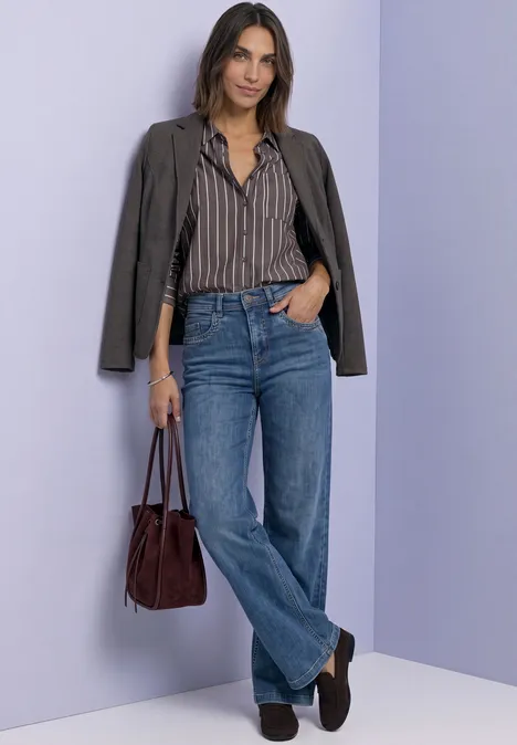 Frau im gestreiften Hemd, Blazer, Blue-Jeans und dunkelroter Tasche vor fliederfarbener Wand.