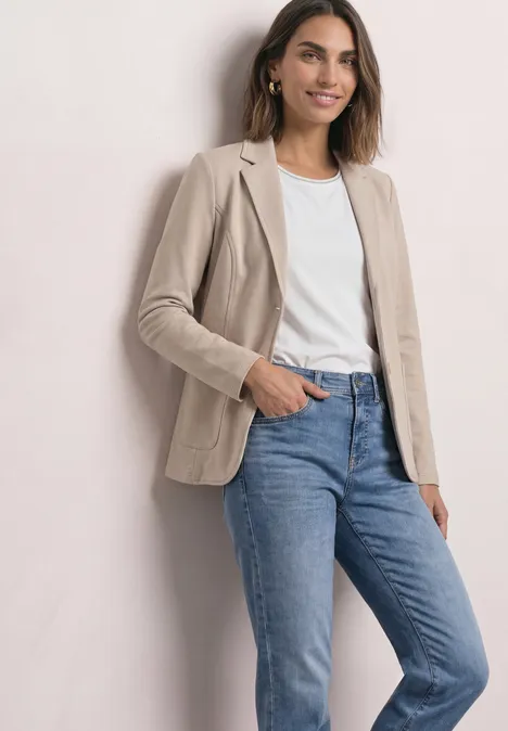 Frau in beigem Blazer, weißem T-Shirt und blauen Jeans vor hellem Hintergrund.
