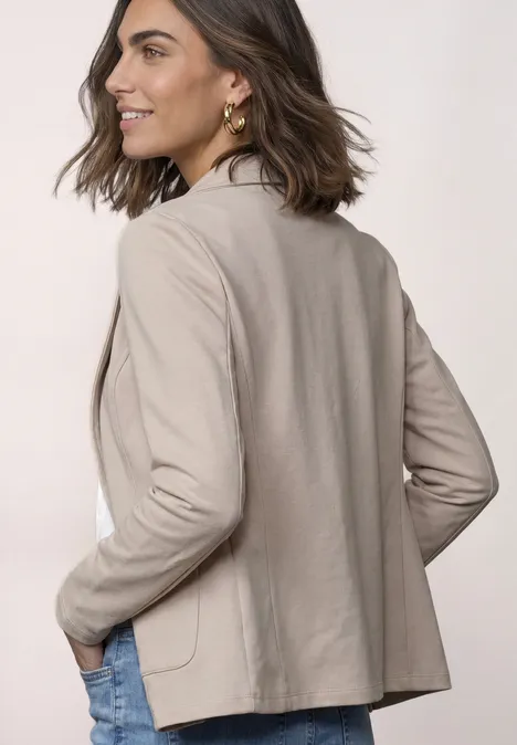 Frau in beige Blazer schaut über die Schulter, Hände in den Taschen, leicht lächelnd.