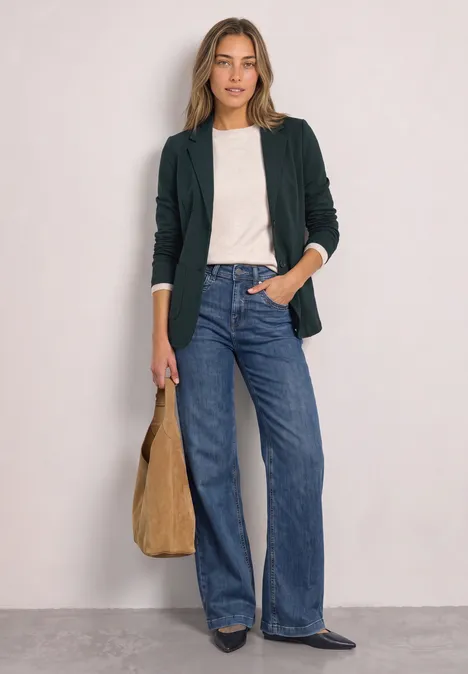 Frau trägt weißen Pullover, dunkelgrünen Blazer, blaue Jeans und hält braune Tasche
