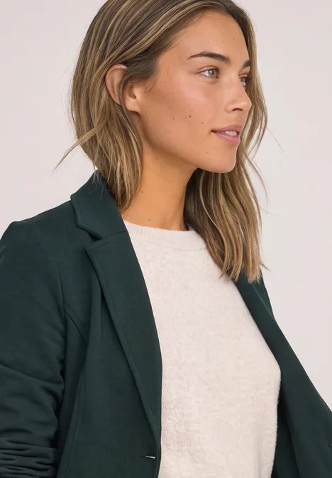 Frau mit kurzem hellbraunem Haar in dunkelgrünem Blazer und weißem Shirt, seitlich vor hellem Hintergrund.