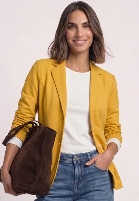 Eine lächelnde Frau in gelbem Blazer, weißem Shirt, Jeans und brauner Wildledertasche.