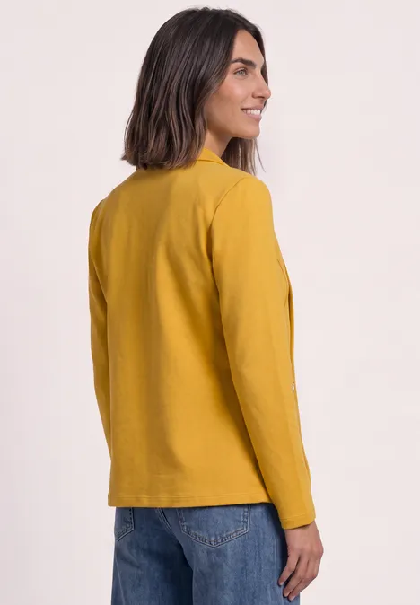 Frau im senfgelben Blazer und blauen Jeans, halb seitlich von hinten fotografiert.