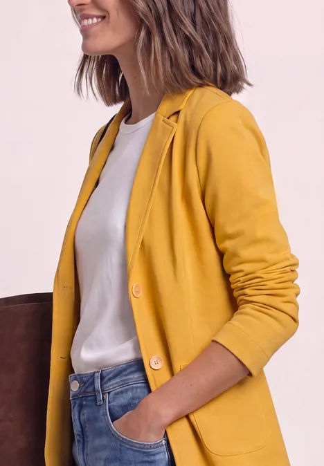 Frau trägt gelben Blazer, weißes T-Shirt und Jeans, eine Hand in der Tasche, lässiger Look.