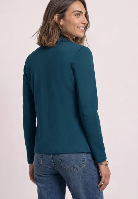 Frau in tealfarbenem Langarmshirt und Jeans blickt lächelnd über die Schulter.