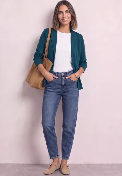 Eine Frau steht entspannt in grünem Blazer, weißem T-Shirt, blauen Jeans, beigen Schuhen und Schultertasche.