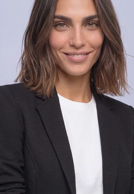 Eine lächelnde Frau mit schulterlangen braunen Haaren, die ein weißes T-Shirt und schwarzen Blazer trägt.