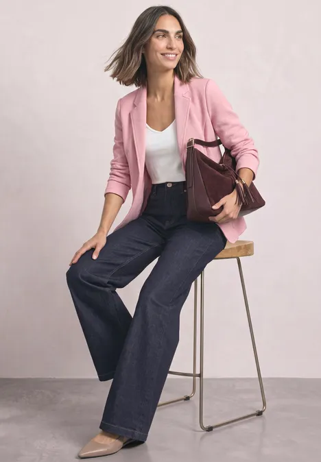 Frau in rosa Blazer, weißem Top und weiten Jeans sitzt auf Hocker und hält bordeauxrote Tasche.