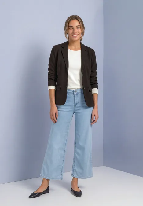 Jeune femme en blazer noir, t-shirt blanc, jean large et chaussures noires, posant devant un mur clair.