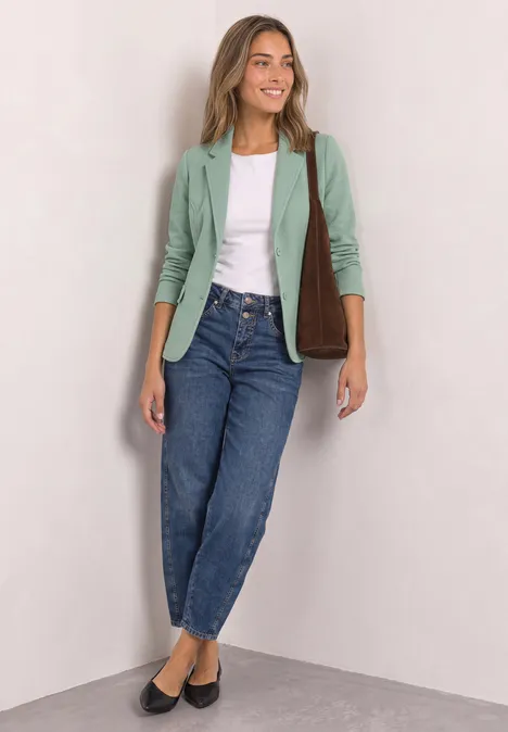 Frau in grünem Blazer, weißem Top, Jeans und schwarzen Ballerinas, mit brauner Schultertasche vor hellem Hintergrund