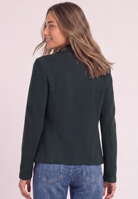 Eine Frau von hinten in einem schwarzen Blazer und blauen Jeans vor neutralem Hintergrund.