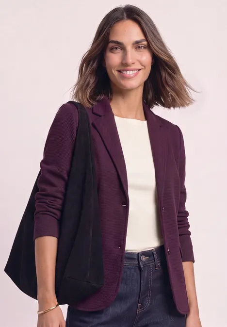Junge Frau lächelt, trägt lila Blazer, weißes Shirt, Jeans und schwarze Tasche.