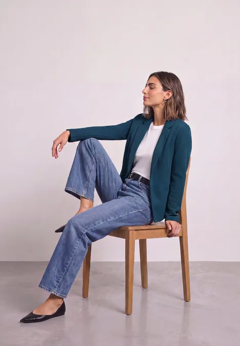 Frau sitzt lässig auf einem Holzstuhl, trägt Jeans, weiße Bluse und grünen Blazer.