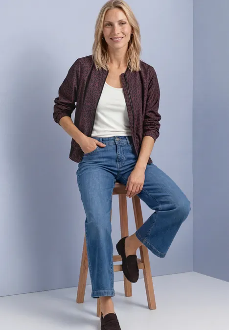 Blonde Frau sitzt lächelnd auf einem Hocker, trägt weißes Top, gemusterte Bomberjacke und blaue Jeans.