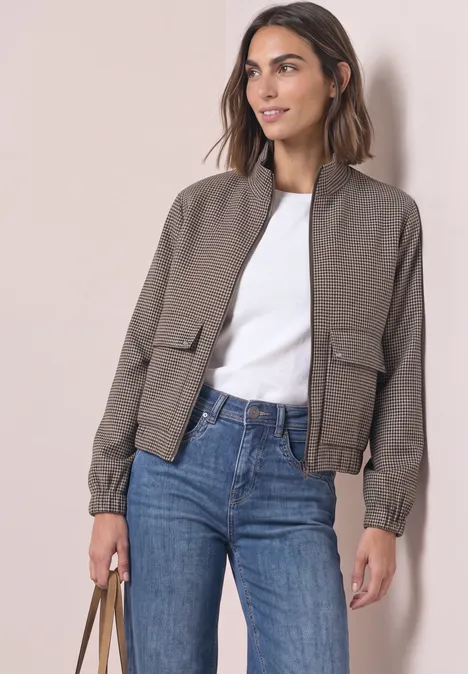 Femme portant une veste bomber taupe, t-shirt blanc et jean, sac à main sur fond clair.