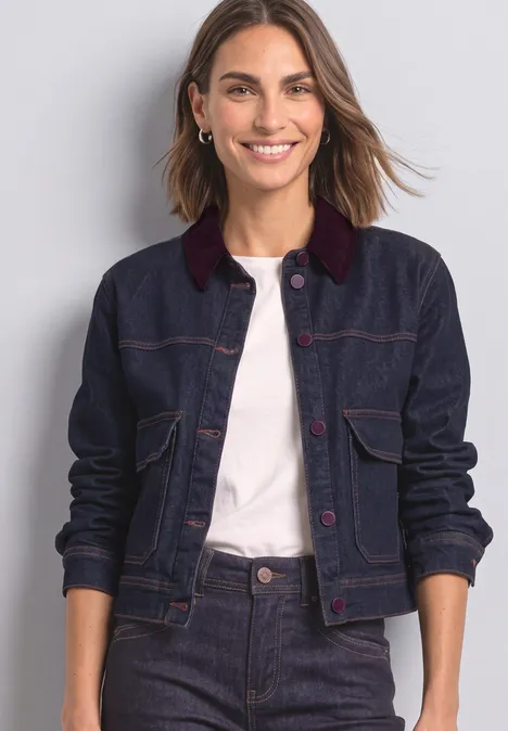Une femme souriante porte une veste en jean bleu foncé au col rouge et un t-shirt blanc sur fond gris clair.