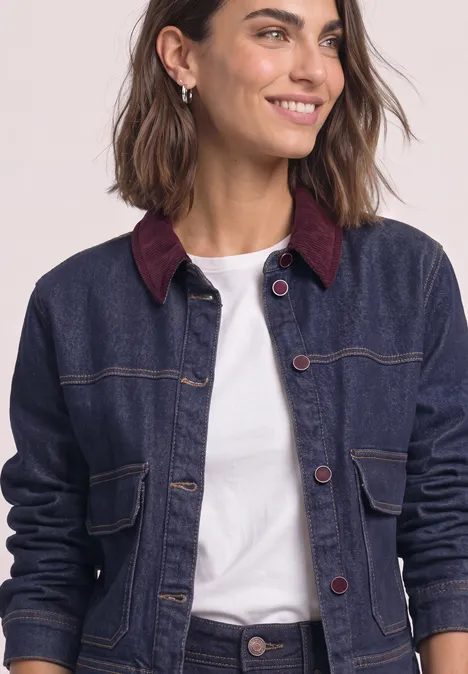 Femme souriante portant une veste en jean bleu foncé à col bordeaux et un T-shirt blanc sur fond clair.