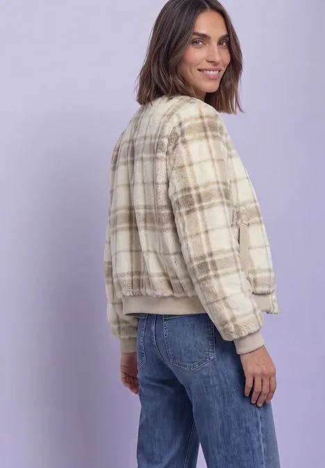 Une femme porte une veste bomber à carreaux et un jean, regarde par-dessus l’épaule en souriant sur fond pastel.