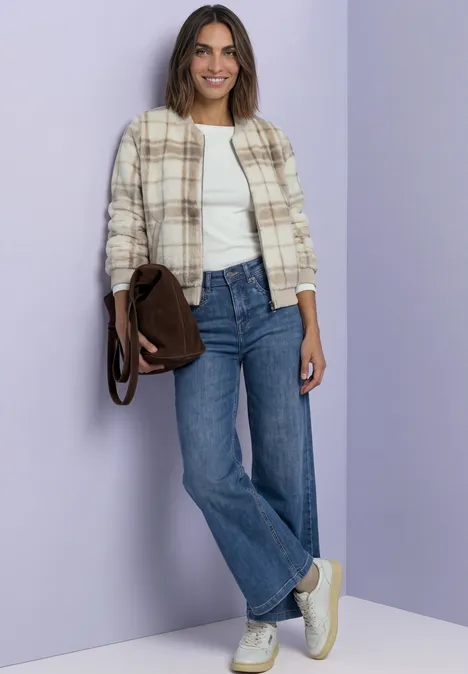 Une femme en jean, top blanc et veste à carreaux, posant décontractée avec un sac marron.